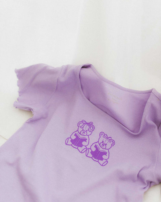 The Book Bears Baby Tee features a design with two teddy bears each holding a book. The t-shirt is a light purple color with a deep purple bookish design and lettuce hem trim.