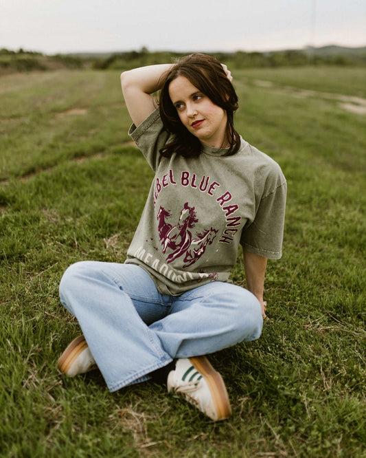 Rebel Blue Ranch Mustangs Tee. A woman wears a mineral wash olive green boxy oversized Rebel Blue Ranch tee with a horses design based on the cowboy romance book series by Lyla Sage that includes Done and Dusted, Swift and Saddled, Lost and Lassoed, and Wild and Wrangled. Meadowlark bookish merch. Lyla Sage merch.