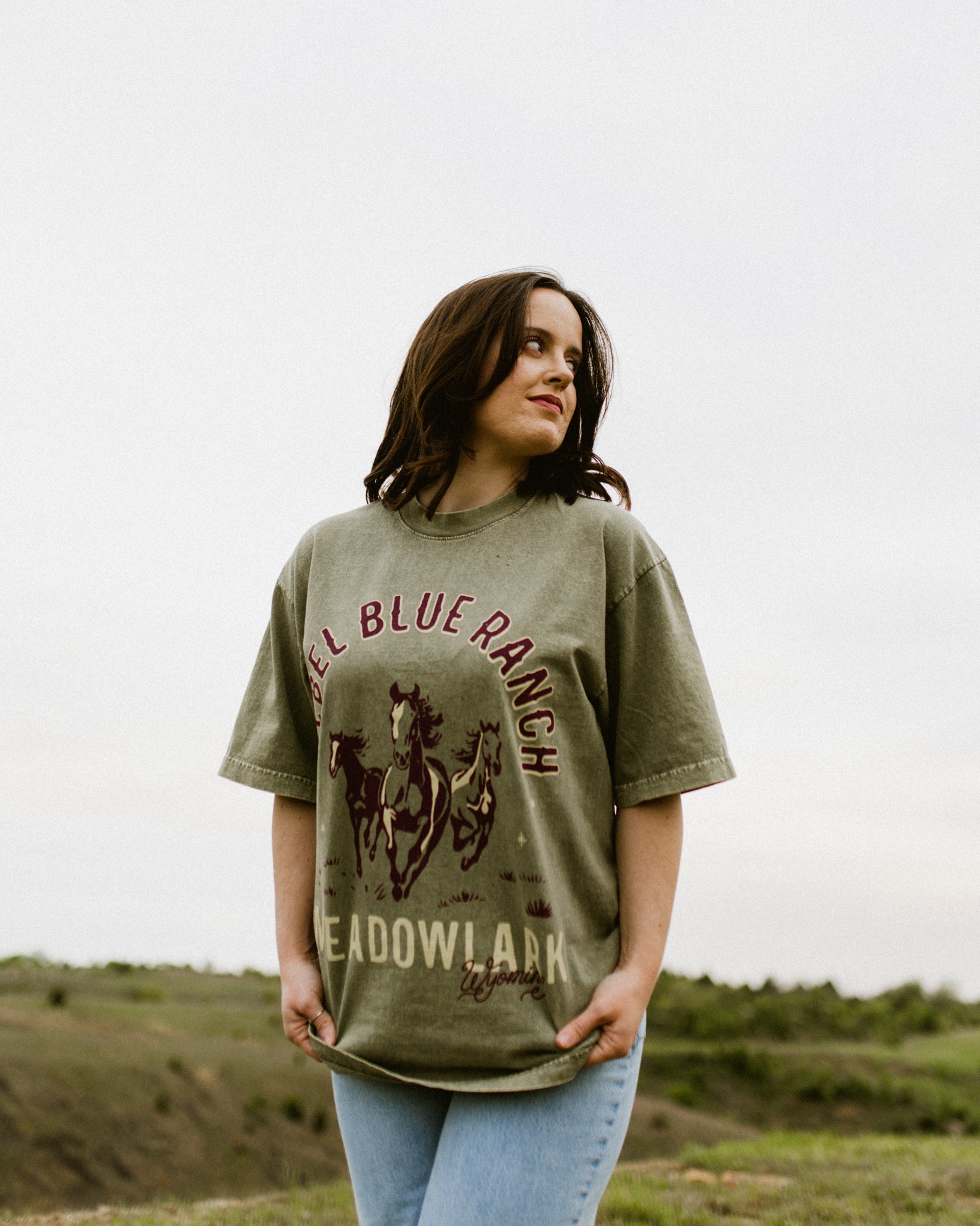 Rebel Blue Ranch Mustangs Tee. A woman wears a mineral wash olive green boxy oversized Rebel Blue Ranch tee with a horses design based on the cowboy romance book series by Lyla Sage that includes Done and Dusted, Swift and Saddled, Lost and Lassoed, and Wild and Wrangled. Meadowlark bookish merch. Lyla Sage merch.