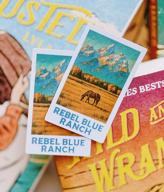 Two Rebel Blue Ranch Polaroid Stickers from 'Rebel Blue Ranch' by Lyla Sage with a horse and landscape design, placed on top of other books.