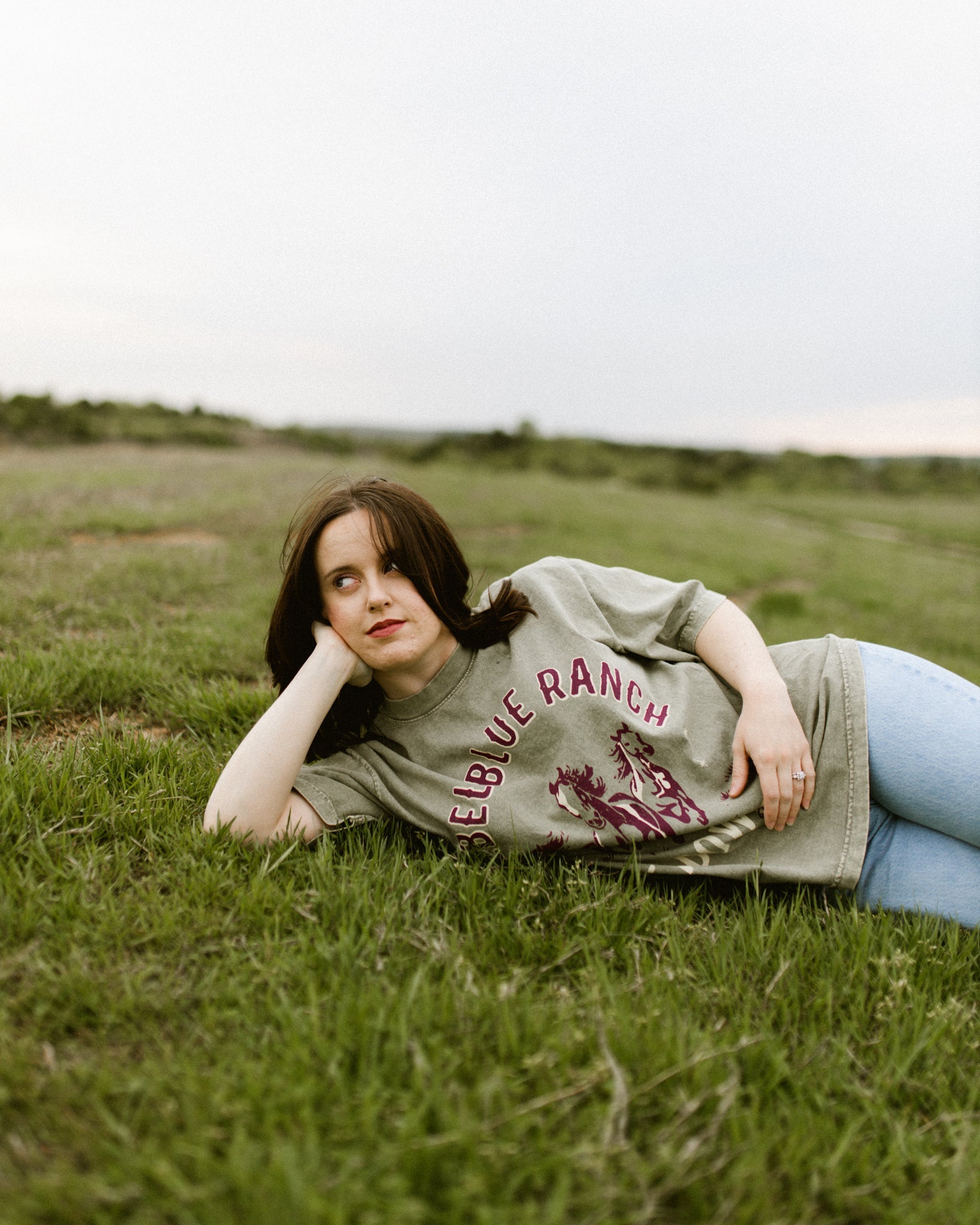 Rebel Blue Ranch Mustangs Tee. A woman wears a mineral wash olive green boxy oversized Rebel Blue Ranch tee with a horses design based on the cowboy romance book series by Lyla Sage that includes Done and Dusted, Swift and Saddled, Lost and Lassoed, and Wild and Wrangled. Meadowlark bookish merch. Lyla Sage merch.
