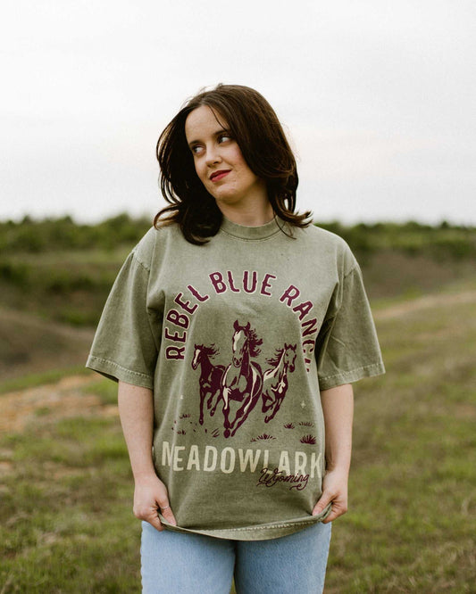 Rebel Blue Ranch Mustangs Tee. A woman wears a mineral wash olive green boxy oversized Rebel Blue Ranch tee with a horses design based on the cowboy romance book series by Lyla Sage that includes Done and Dusted, Swift and Saddled, Lost and Lassoed, and Wild and Wrangled. Meadowlark bookish merch. Lyla Sage merch.