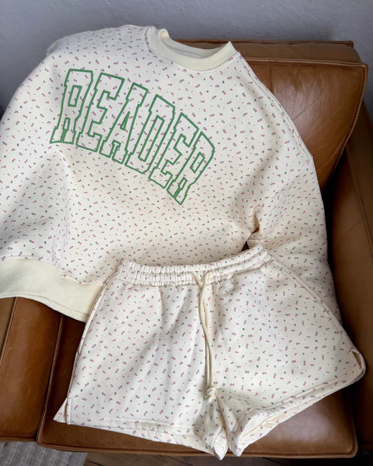 The Reader Floral Crewneck from Next Chapter features a floral patterned fabric with a green puff print 'Reader' design on the front. Pictured with the matching floral shorts. 