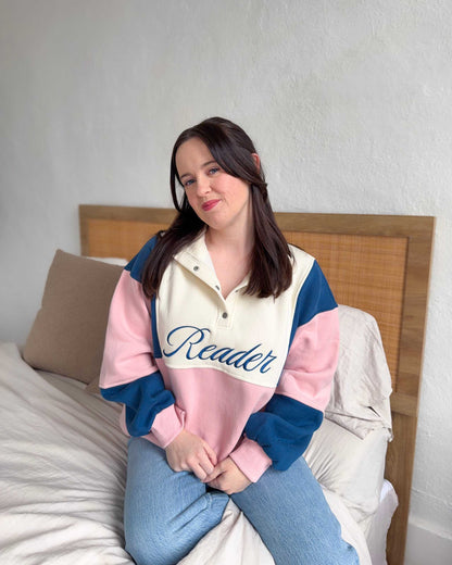 The Reader Colorblock Pullover from Next Chapter is oversized, with a quarter button-up style. This variant is navy, pink, and ivory and has the word 'Reader' embroidered across the front. Next Chapter offers high quality bookish merch and apparel. 