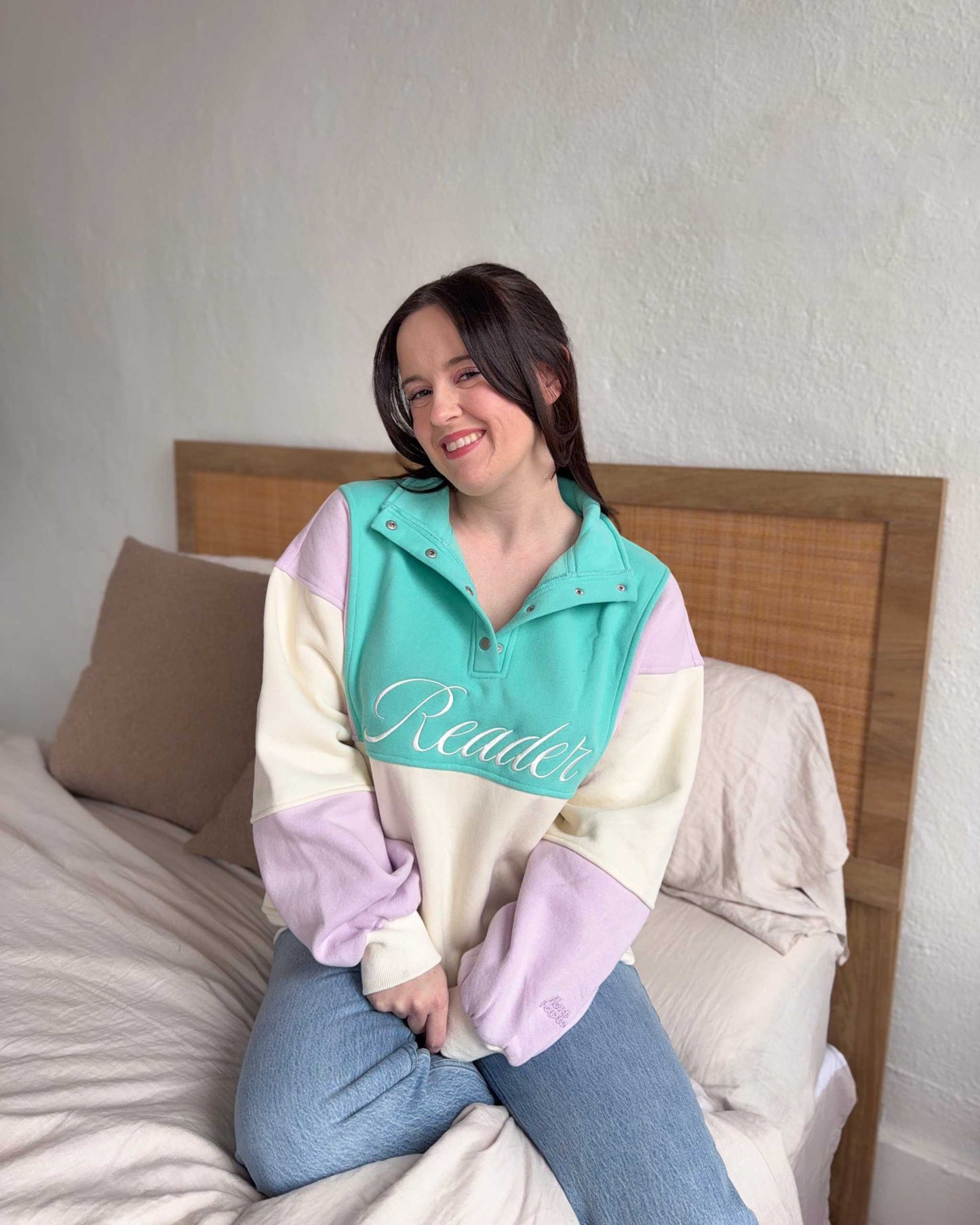 The Reader Colorblock Pullover from Next Chapter is oversized, with a quarter button-up style. This variant is lilac, teal, and ivory and has the word 'Reader' embroidered across the front. Next Chapter offers high quality bookish merch and apparel. 