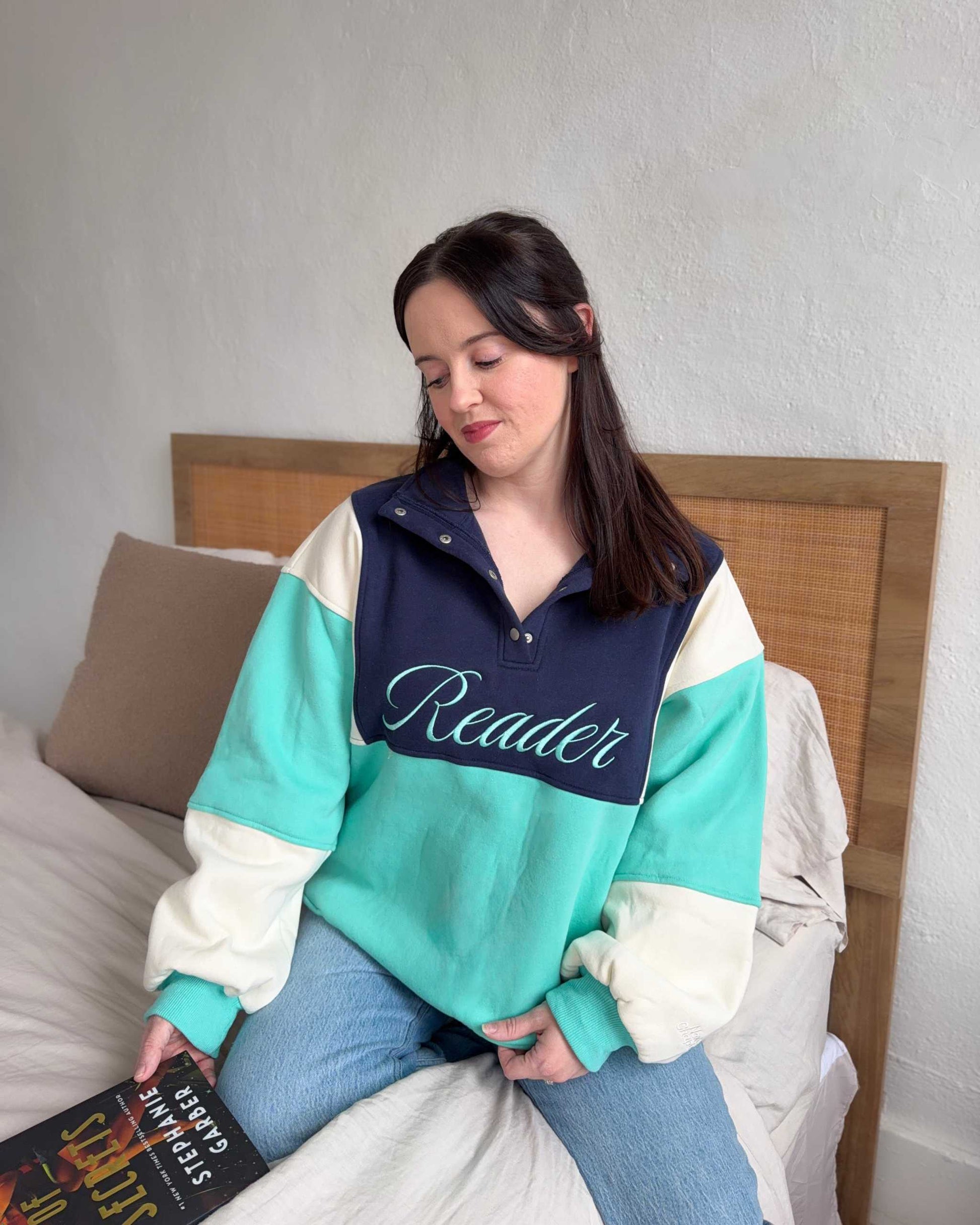 The Reader Colorblock Pullover from Next Chapter is oversized, with a quarter button-up style. This variant is navy, teal, and ivory and has the word 'Reader' embroidered across the front. Next Chapter offers high quality bookish merch and apparel. 