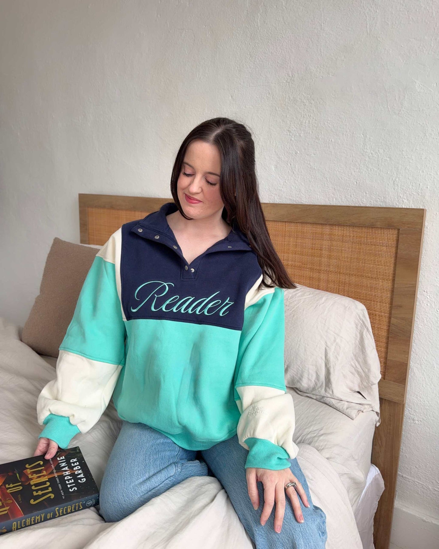 The Reader Colorblock Pullover from Next Chapter is oversized, with a quarter button-up style. This variant is navy, teal, and ivory and has the word 'Reader' embroidered across the front. Next Chapter offers high quality bookish merch and apparel. 