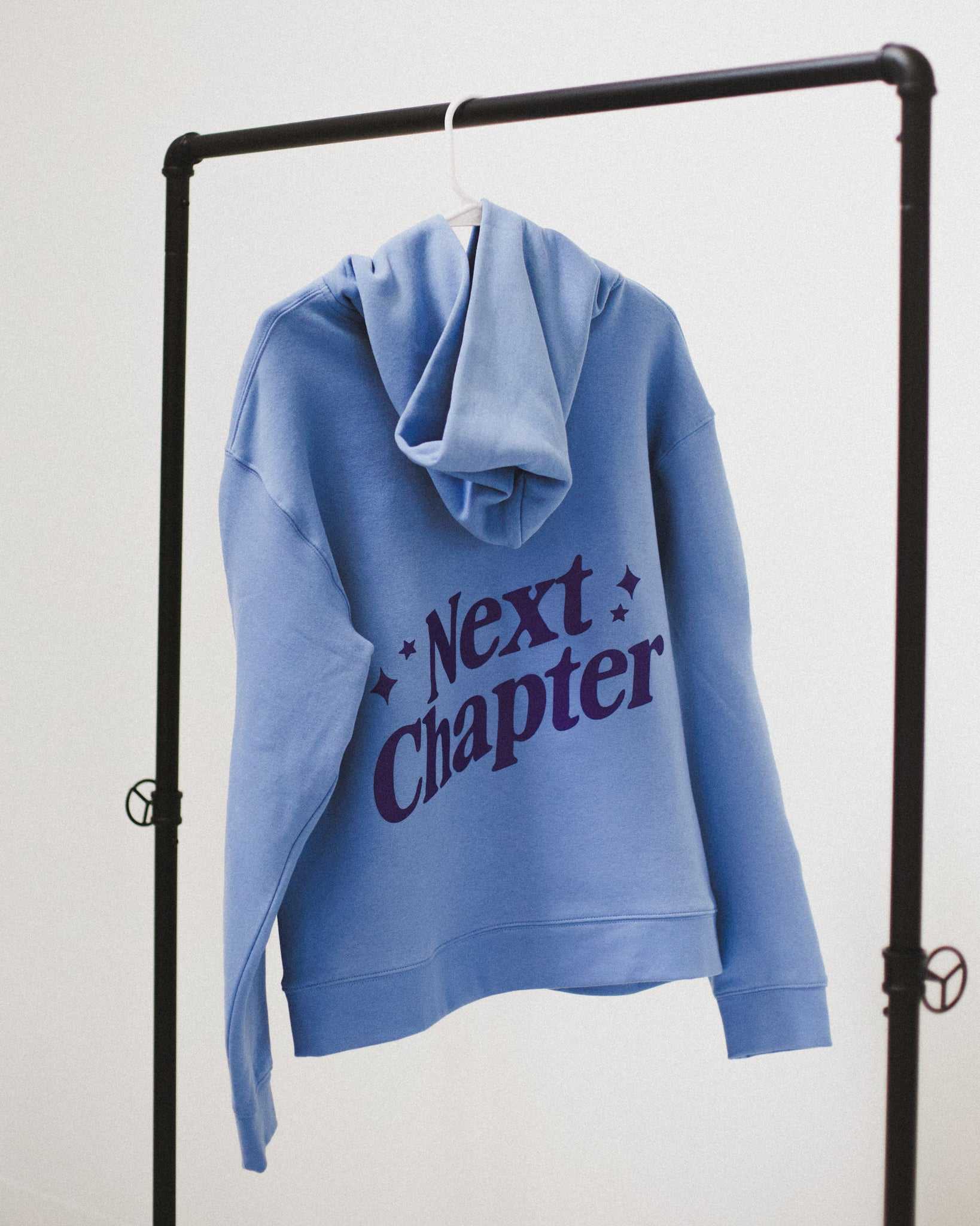 Blue Next Chapter hoodie with 'Next Chapter' puff print text on a black rack against a white background. Next Chapter is a bookish small business that offers officially licensed book merch, including Rachel Gillig merch and Lyla Sage merch.