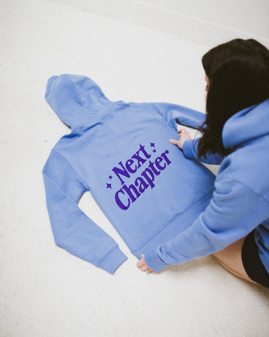 Person wearing a blue hoodie with 'Next Chapter' text on a light background. Next Chapter offers officially licensed book merch, such as Rachel Gillig merch and Lyla Sage merch.