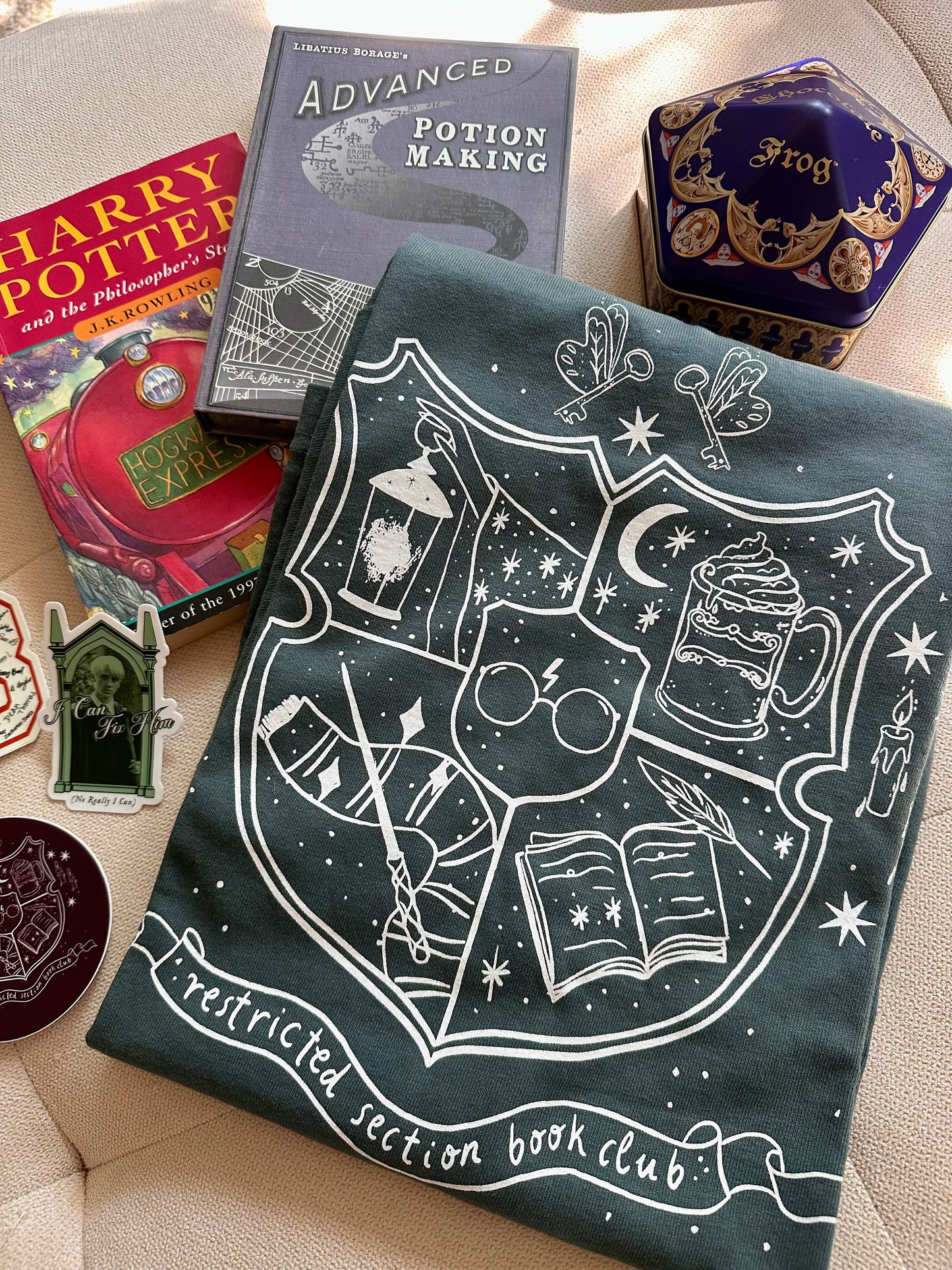 A spruce green long sleeve garment dyed t-shirt is displayed on a flat surface surrounded by Harry Potter books and a chocolate frog tin. The shirt says "restricted section book club" with imagery related to Harry Potter, such as a wand, scarf, glasses, and a book.