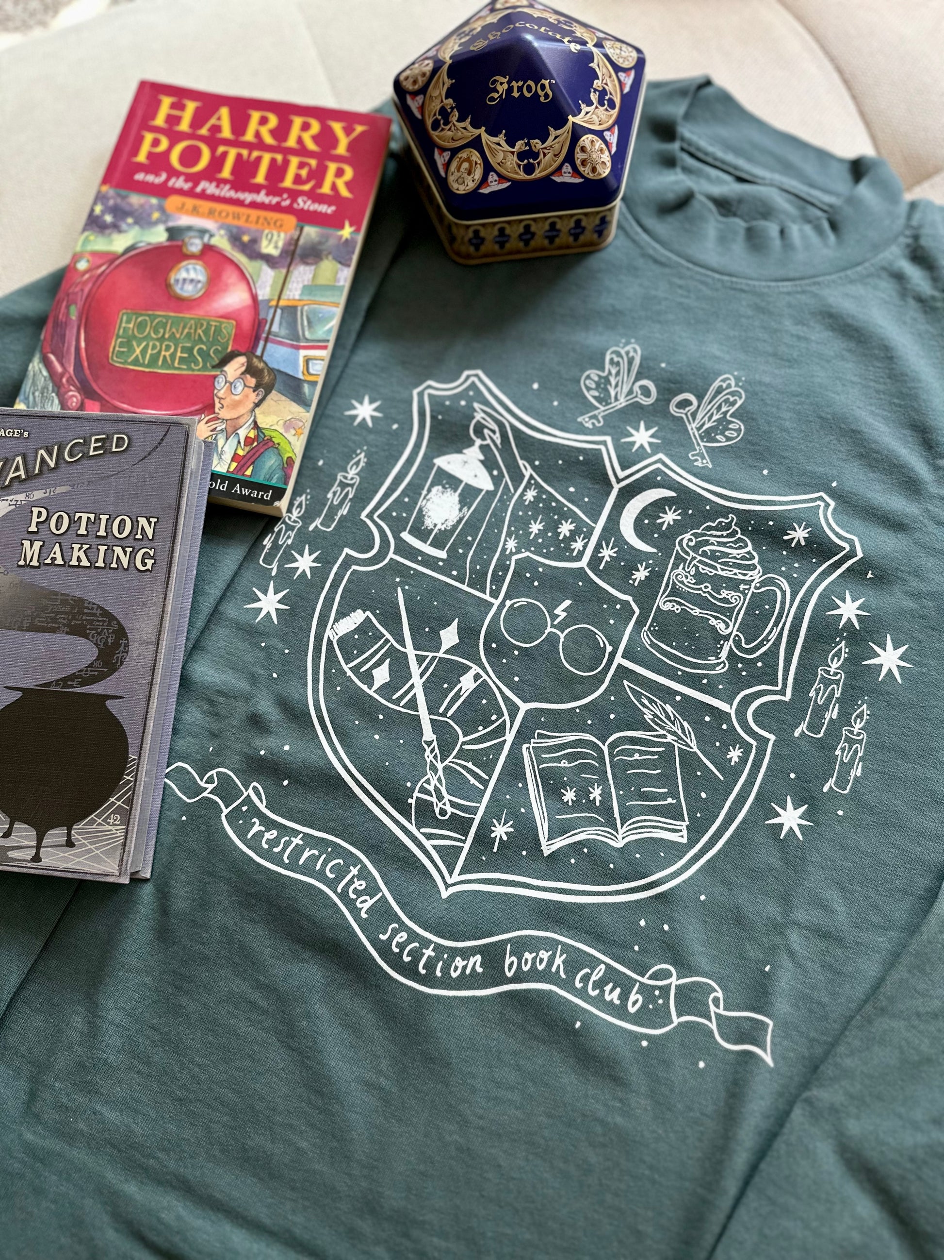 A spruce green long sleeve garment dyed t-shirt is displayed on a flat surface. The shirt says "restricted section book club" with imagery related to Harry Potter, such as a wand, scarf, glasses, butterbeer, and a book.