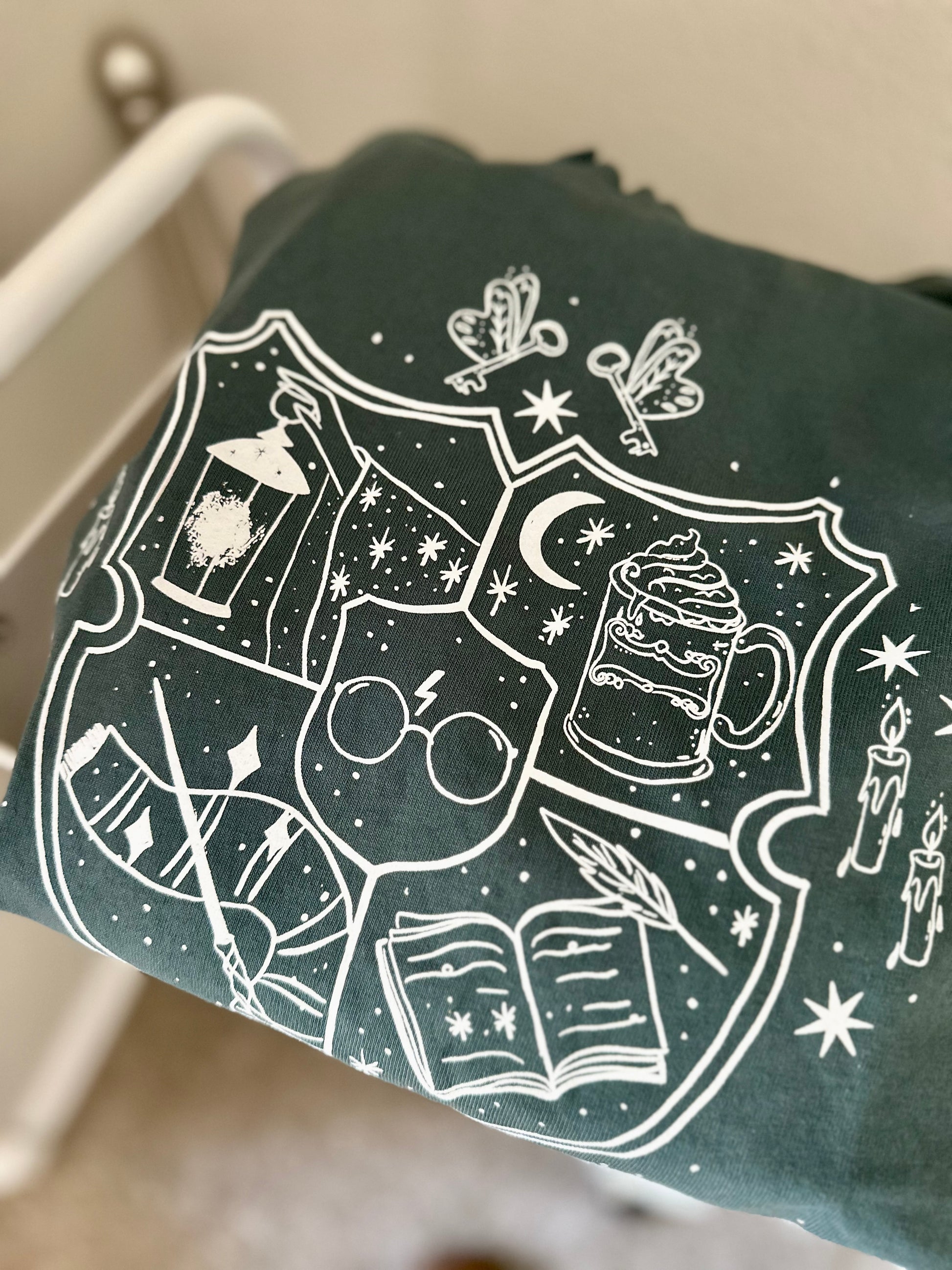 A spruce green long sleeve garment dyed t-shirt is displayed on a flat surface. The shirt says "restricted section book club" with imagery related to Harry Potter, such as a wand, scarf, glasses, butterbeer, and a book.