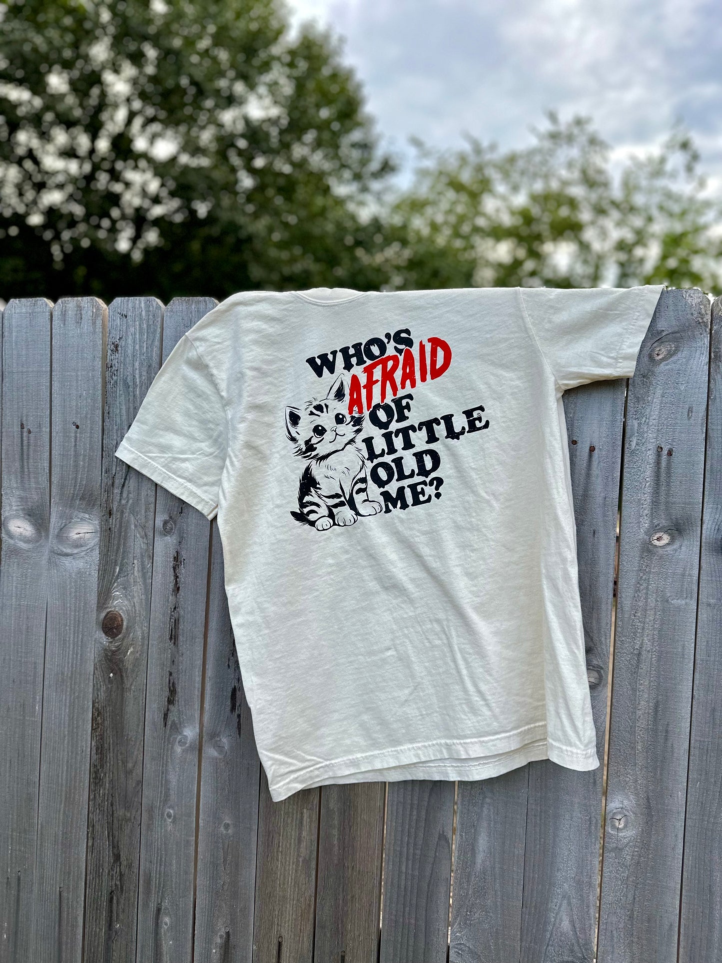 A-shirt with a Taylor Swift lyric is draped over an outdoor fence. The shirt has a kitten on it with the quote "who's afraid of little old me?" The shirt is ivory with a black and red design. 