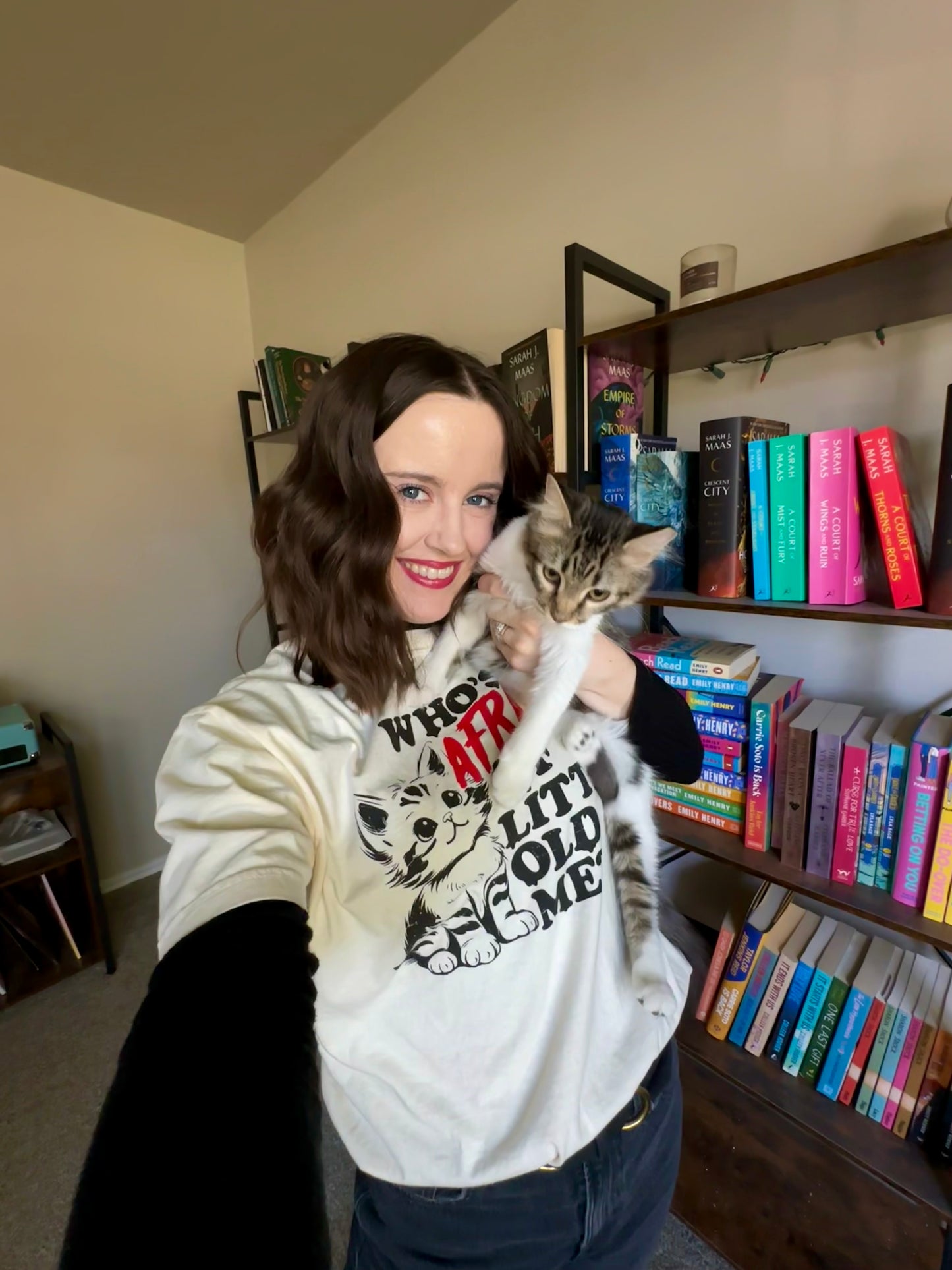 A woman is wearing a shirt with a Taylor Swift lyric while holding a cat. The shirt has a kitten on it with the quote "who's afraid of little old me?" The shirt is ivory with a black and red design. 