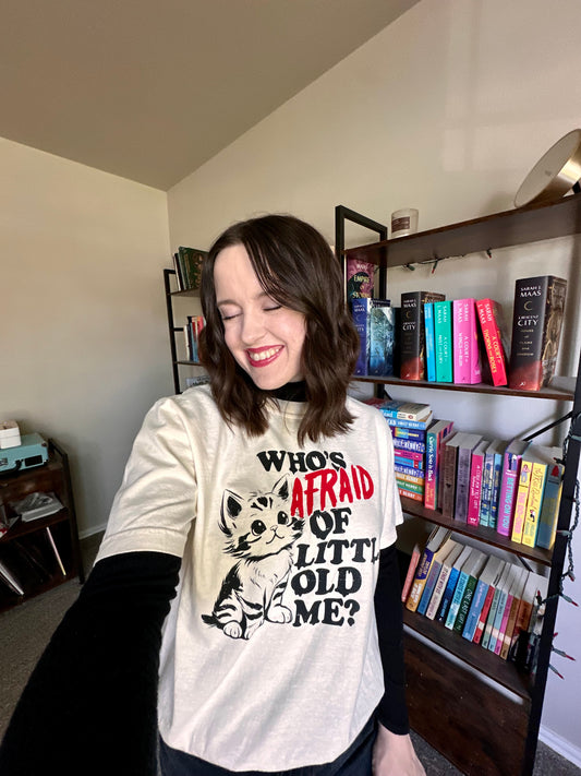 A person stands in front of a bookshelf wearing a t-shirt with a Taylor Swift lyric. The shirt has a kitten on it with the quote "who's afraid of little old me?" The shirt is ivory with a black and red design. 