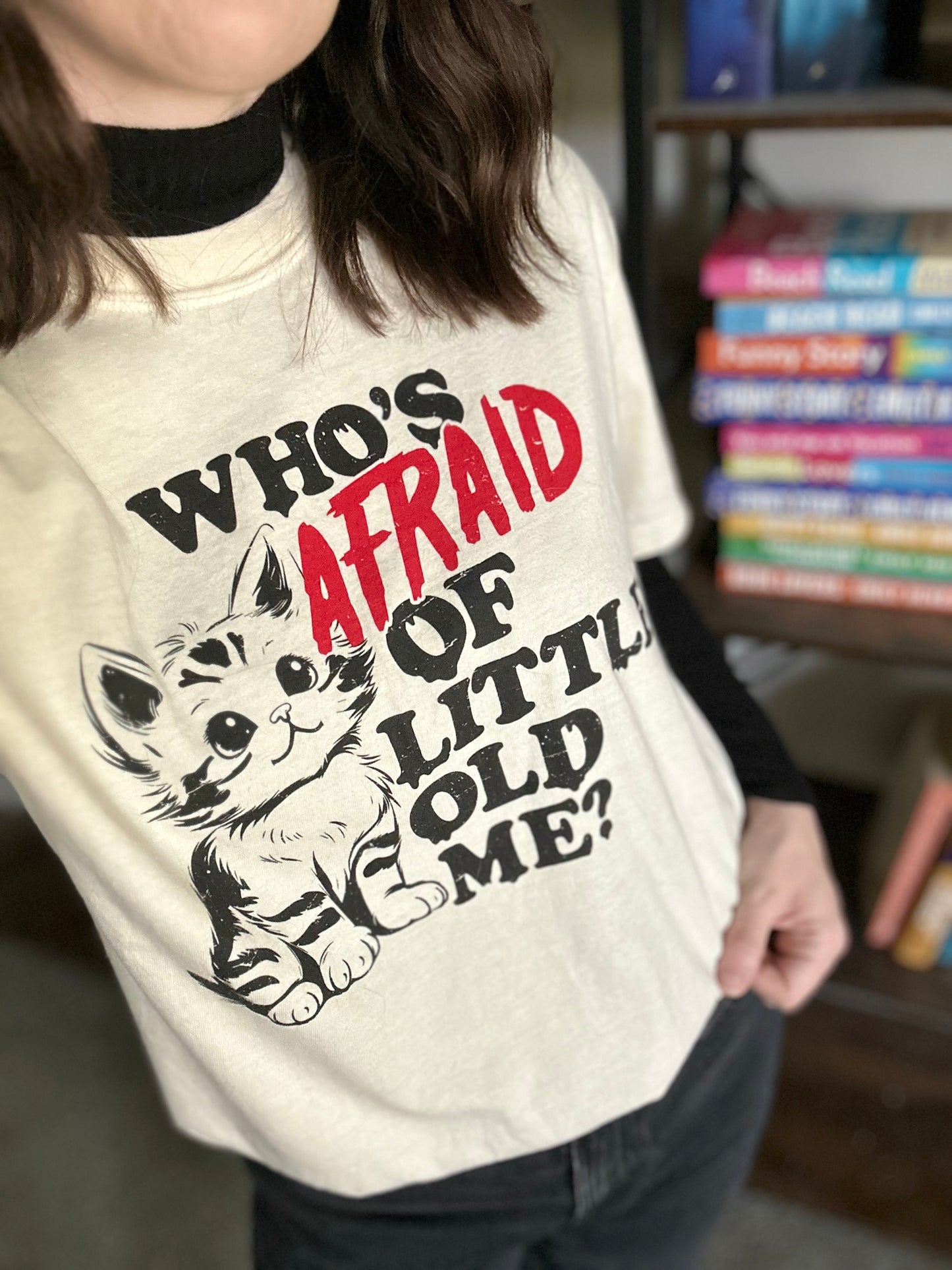 A person stands in front of a bookshelf wearing a t-shirt with a Taylor Swift lyric. The shirt has a kitten on it with the quote "who's afraid of little old me?" The shirt is ivory with a black and red design. 