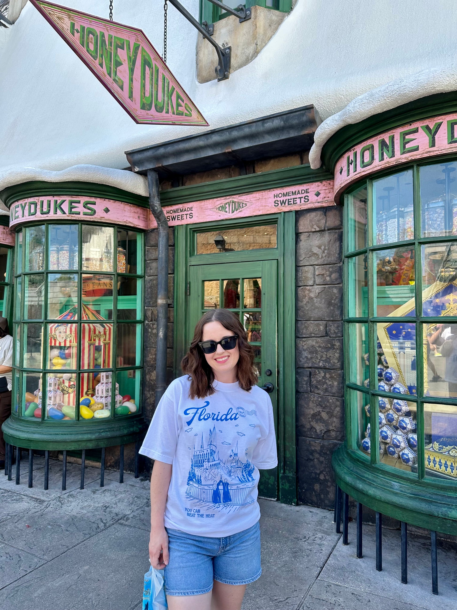 A woman stands outside Honeydukes wearing a t-shirt that depicts the Wizarding World of Harry Potter theme park. The t-shirt also references Taylor Swift's song "Florida!!!" as well as the opening date of the park. 