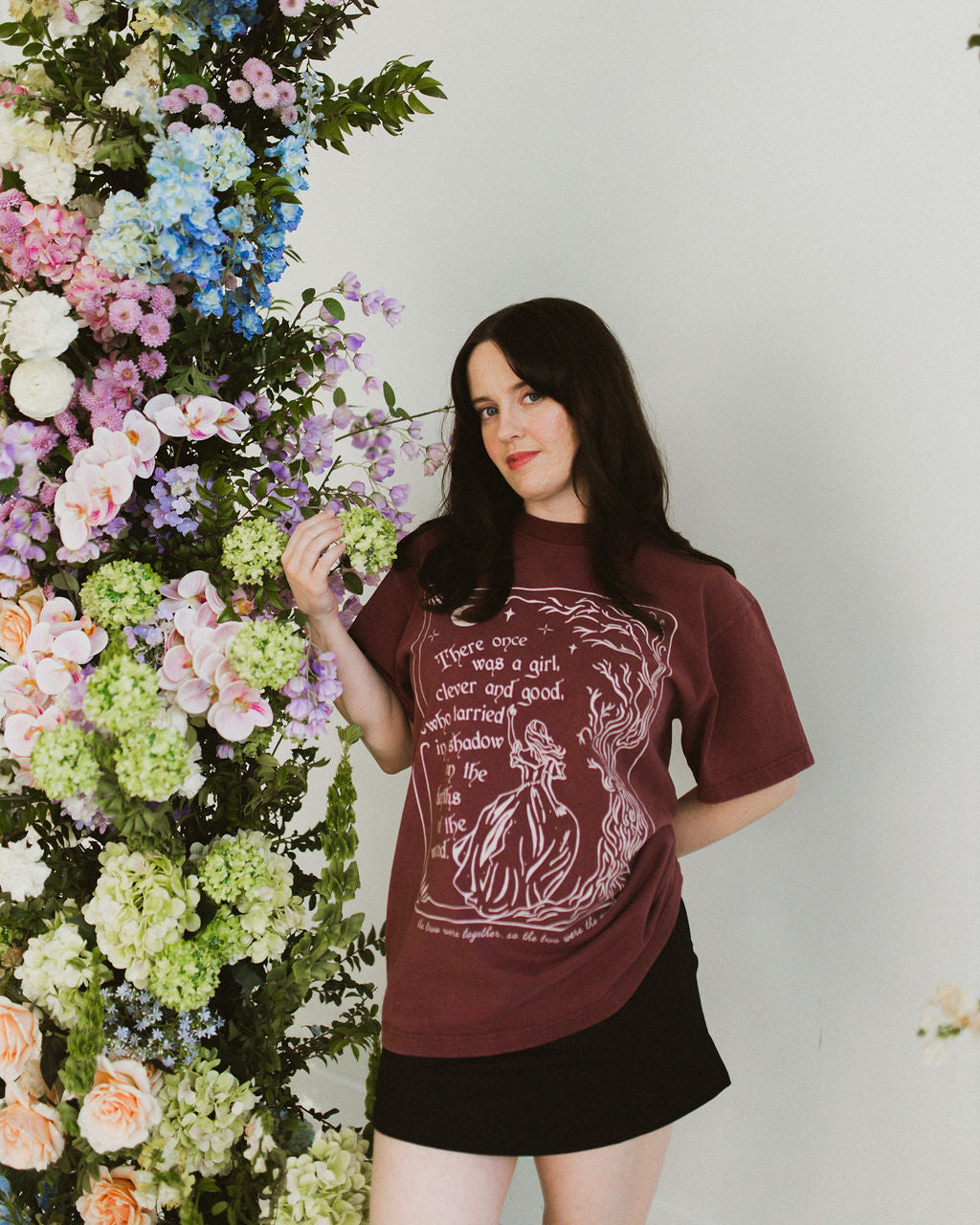 The One Dark Window Tee from Next Chapter is officially licensed by Rachel Gillig and inspired by the Shepherd King Duology. Next Chapter is an official retailer of Rachel Gillig licensed merchandise. This image shows a model wearing the One Dark Window Tee in the maroon color option.