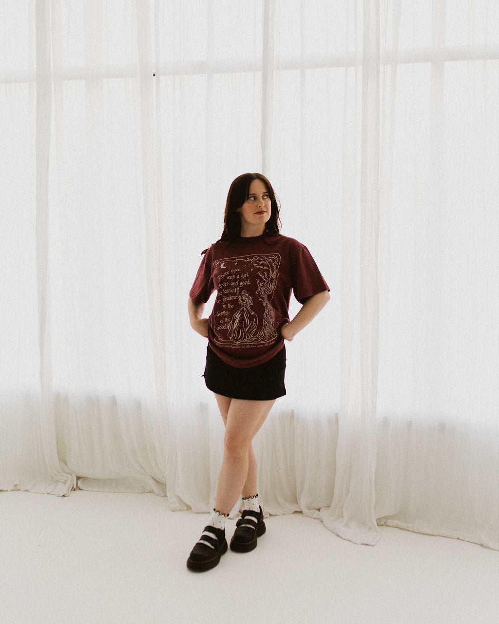 The One Dark Window Tee from Next Chapter is officially licensed by Rachel Gillig and inspired by the Shepherd King Duology. Next Chapter is an official retailer of Rachel Gillig licensed merchandise. This image shows a model wearing the One Dark Window Tee in the maroon color option.