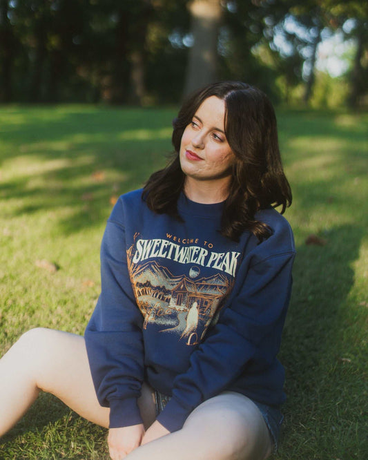 Woman wearing a blue sweatshirt with a graphic design sitting on grass. The Sweetwater Peak crewneck design is inspired by the Lyla Sage book, Soul Searching, which is from her new Sweetwater Peak book series. Merch for book lovers. Officially licensed by Lyla Sage.
