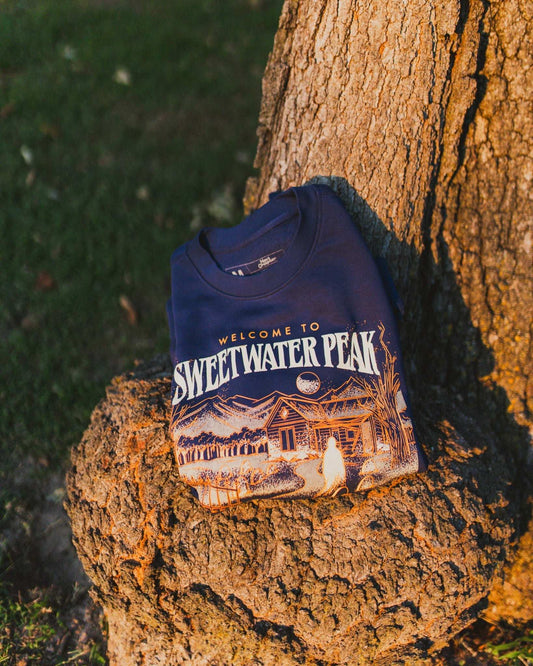 Navy blue sweatshirt with 'Welcome to Sweetwater Peak' print sitting on a tree stump. The Sweetwater Peak crewneck sweatshirt design is inspired by the new book, Soul Searching, which is from the Sweetwater Peak book series by Lyla Sage. Officially licensed by Lyla Sage.