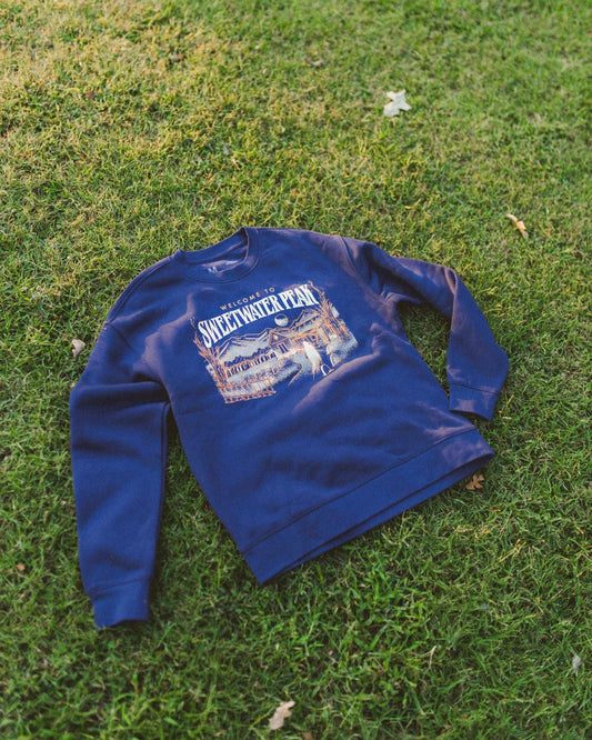 Blue sweatshirt with graphic design on grass. The Sweetwater Peak crewneck design is inspired by the Lyla Sage book, Soul Searching, which is from her new Sweetwater Peak book series. Merch for book lovers. Officially licensed by Lyla Sage.