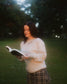Woman wearing oatmeal beige Reader Embroidered Sweater with golden brown text outdoors in autumn. The sweater reads "Reader" in embroidery around the collar. 