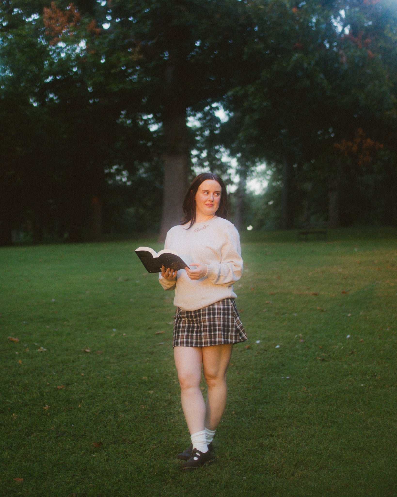 Woman wearing oatmeal beige Reader Embroidered Sweater with golden brown text outdoors in autumn. The sweater reads "Reader" in embroidery around the collar. 