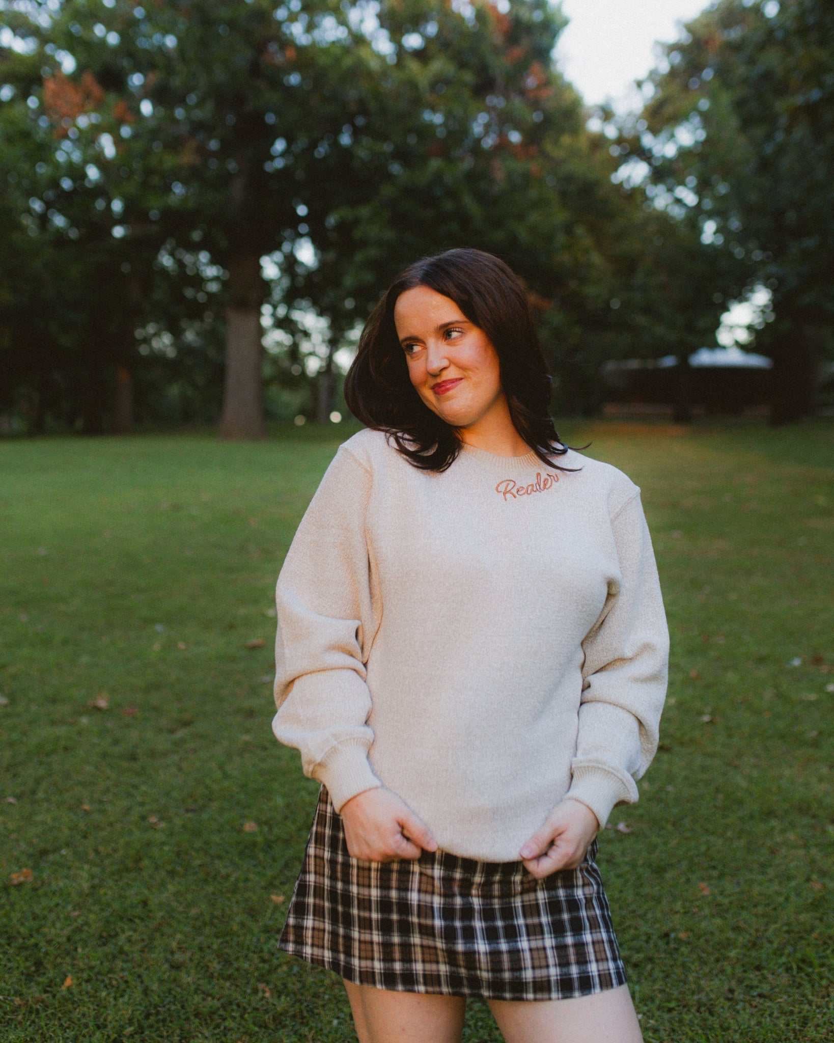 Woman wearing oatmeal beige Reader Embroidered Sweater with golden brown text outdoors in autumn. The sweater reads "Reader" in embroidery around the collar. 
