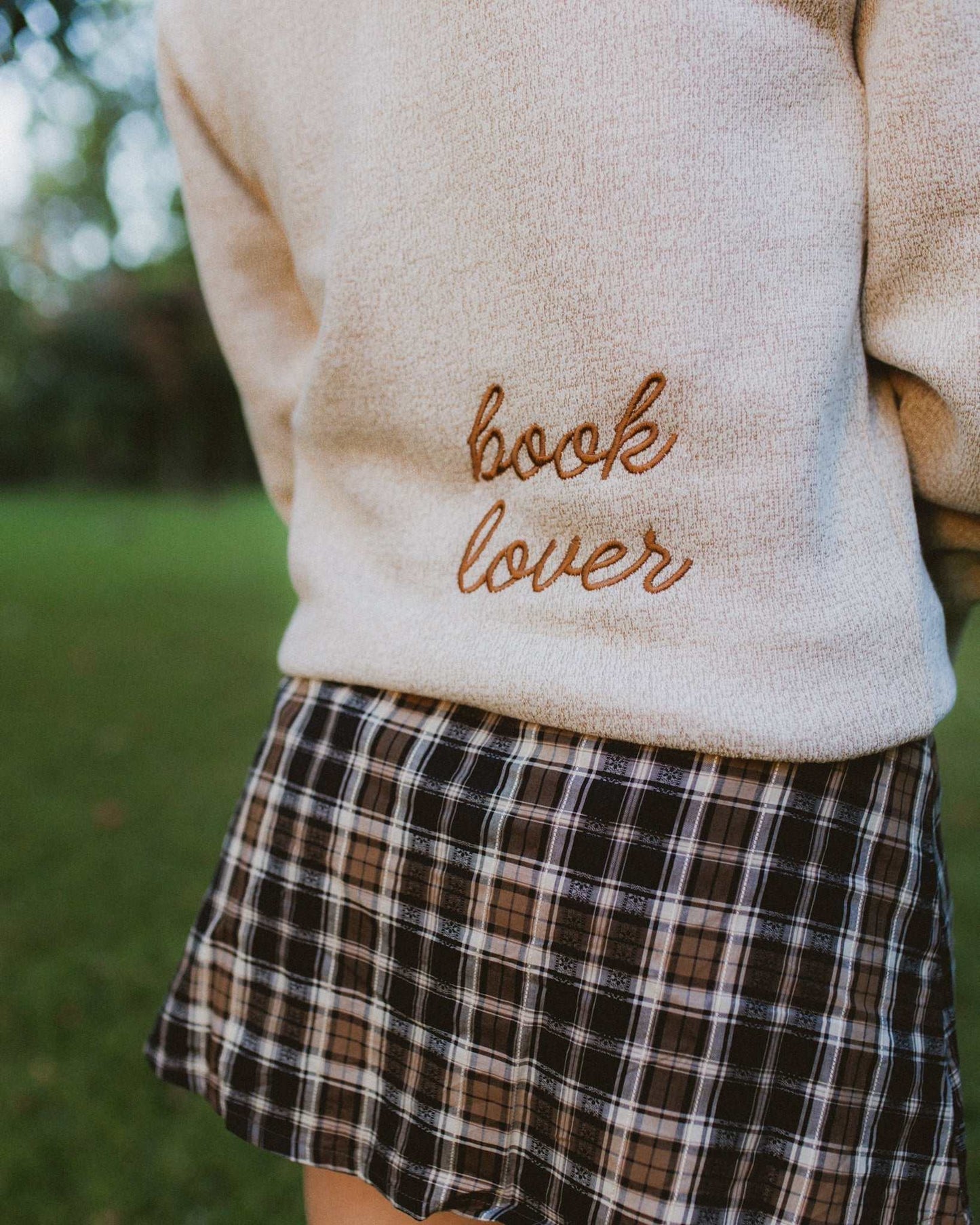 Woman wearing oatmeal beige Reader Embroidered Sweater with golden brown text outdoors in autumn. The sweater reads "book lover" in embroidery on the back of the sweater.