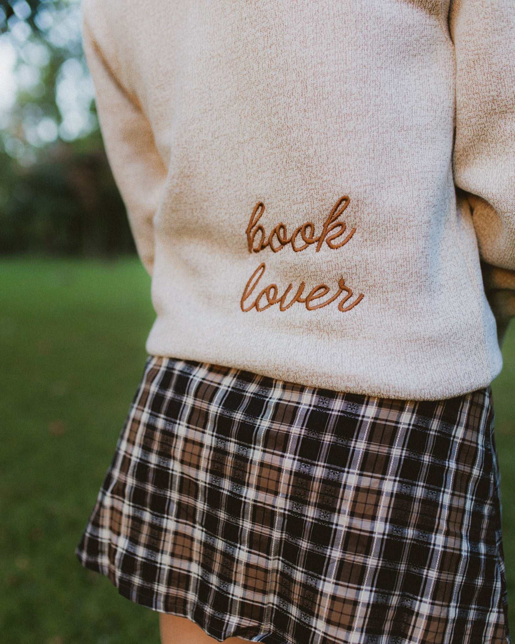 Woman wearing oatmeal beige Reader Embroidered Sweater with golden brown text outdoors in autumn. The sweater reads "book lover" in embroidery on the back of the sweater.