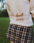 Woman wearing oatmeal beige Reader Embroidered Sweater with golden brown text outdoors in autumn. The sweater reads "book lover" in embroidery on the back of the sweater.