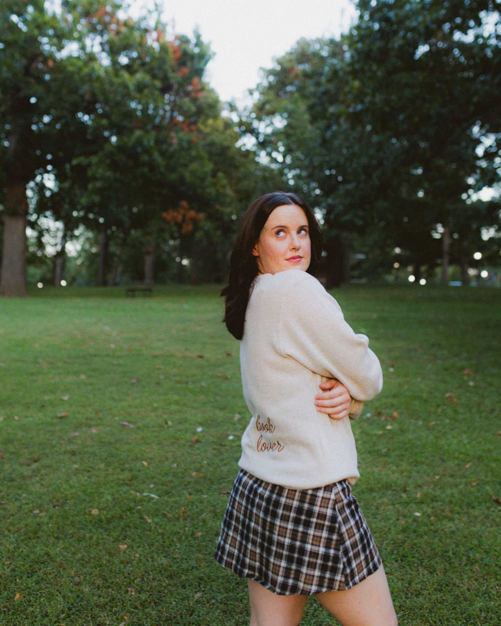 Woman wearing oatmeal beige Reader Embroidered Sweater with golden brown text outdoors in autumn. The sweater reads "book lover" in embroidery on the back of the sweater.