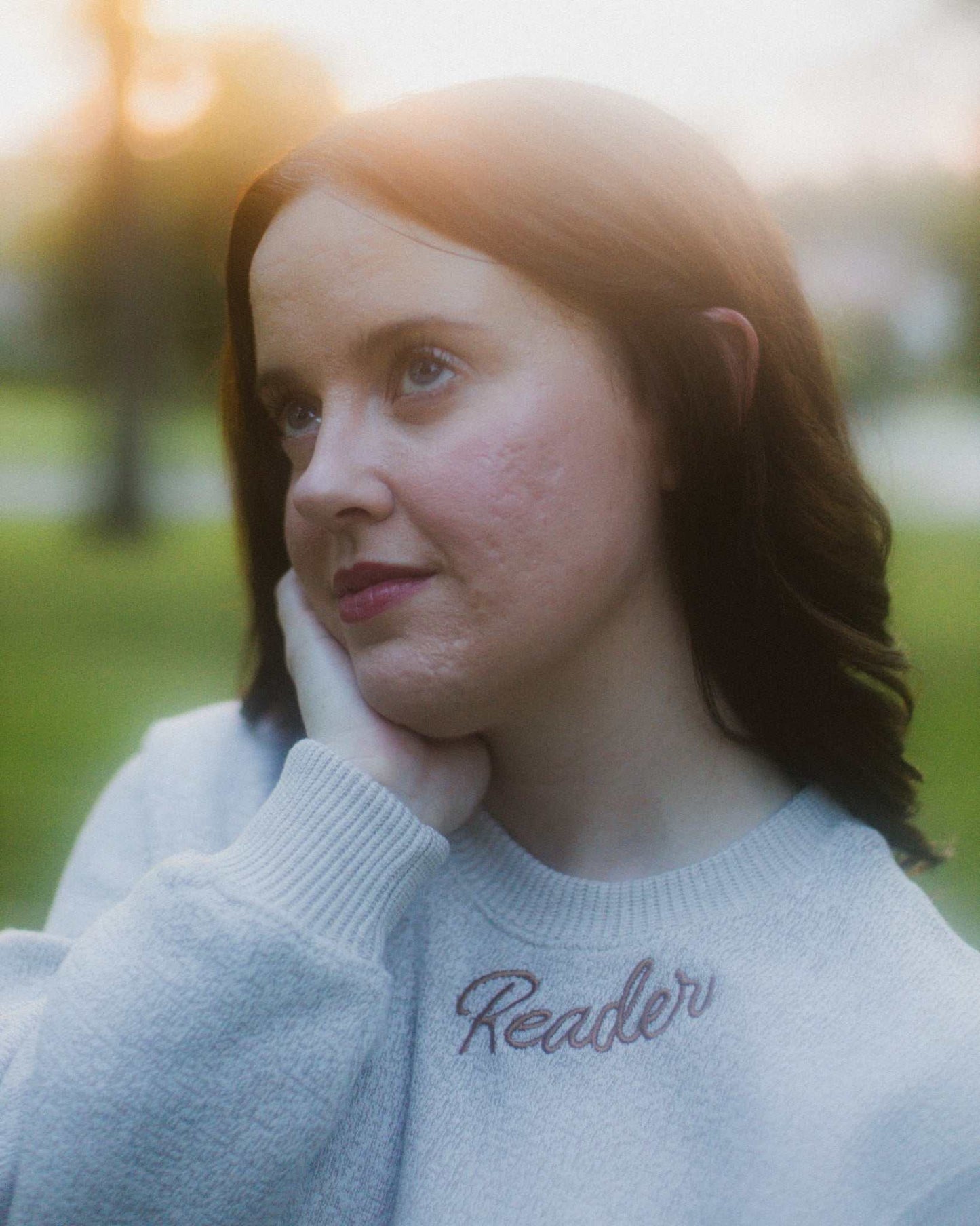 Woman wearing oatmeal beige Reader Embroidered Sweater with golden brown text outdoors in autumn. The sweater reads "Reader" in embroidery around the collar. 