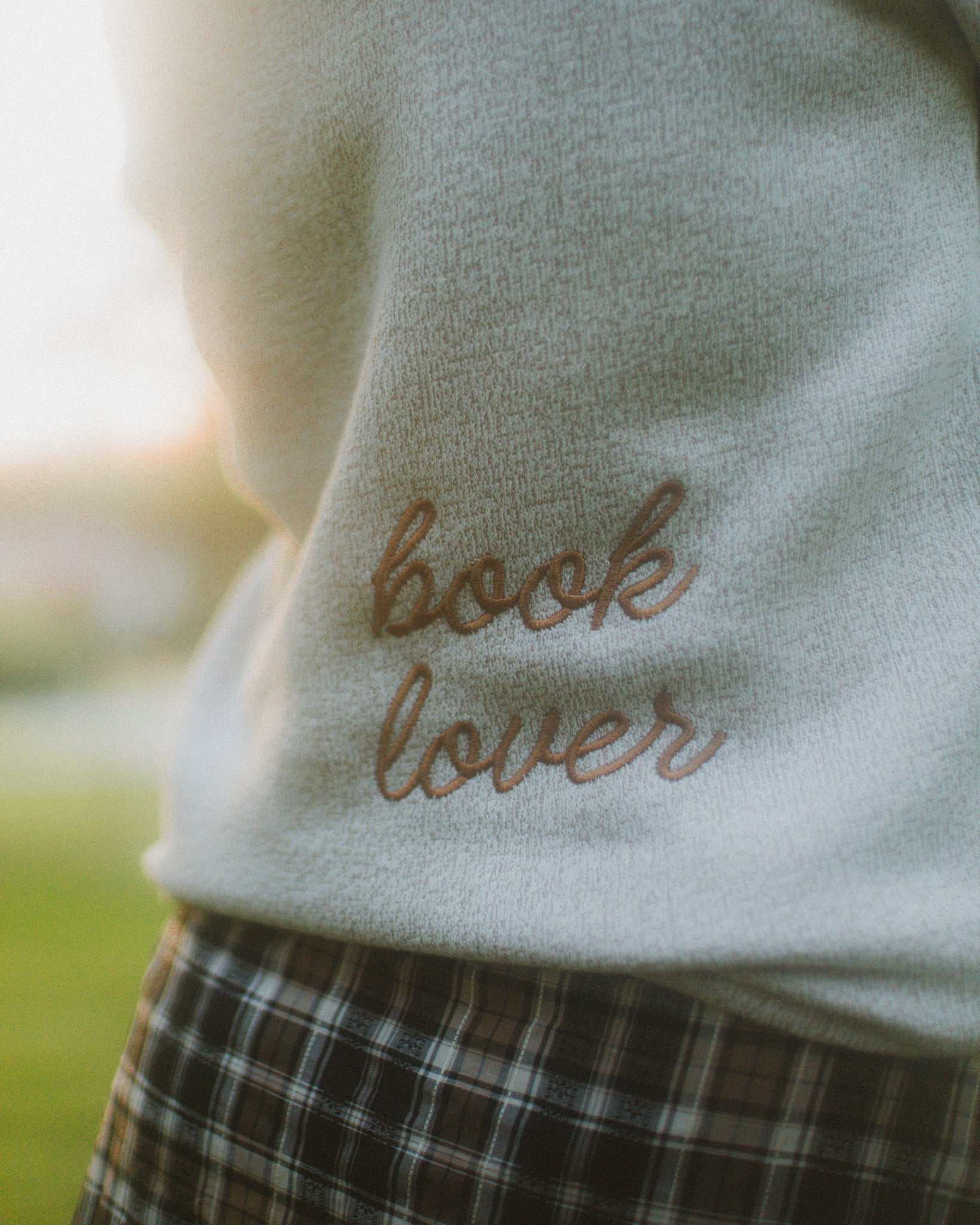 Woman wearing oatmeal beige Reader Embroidered Sweater with golden brown text outdoors in autumn. The sweater reads "book lover" in embroidery on the back of the sweater.