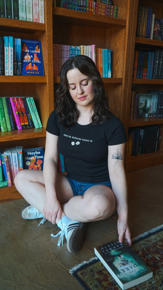 Woman sitting on the floor in front of a bookshelf with books around her. The Thirteen Tee from Throne of Glass by Sarah J. Maas. "Until the darkness claims us" Ironteeth Witches design. 