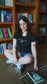Woman sitting on the floor in front of a bookshelf with books around her. The Thirteen Tee from Throne of Glass by Sarah J. Maas. "Until the darkness claims us" Ironteeth Witches design. 