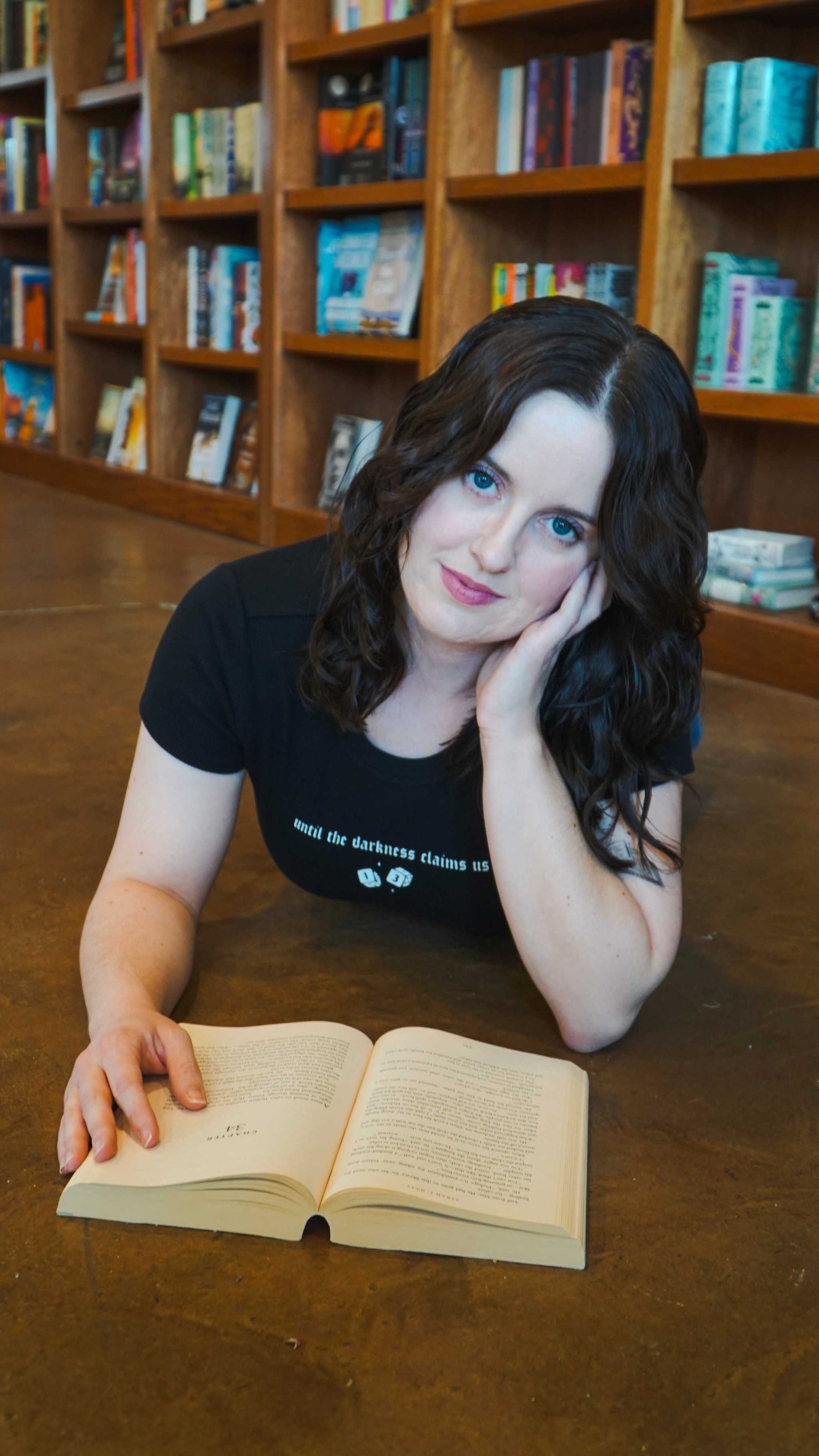 Woman lying on a wooden floor with an open book in a library setting, The Thirteen Tee from Throne of Glass by Sarah J. Maas. "Until the darkness claims us" Ironteeth Witches design. 