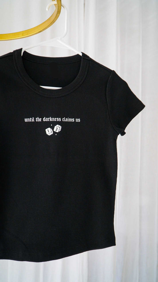 Black t-shirt with white text and graphics on a hanger against a white curtain background. The Thirteen Tee from Throne of Glass by Sarah J. Maas. "Until the darkness claims us" Ironteeth Witches design. 