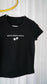 Black t-shirt with white text and graphics on a hanger against a white curtain background. The Thirteen Tee from Throne of Glass by Sarah J. Maas. "Until the darkness claims us" Ironteeth Witches design. 
