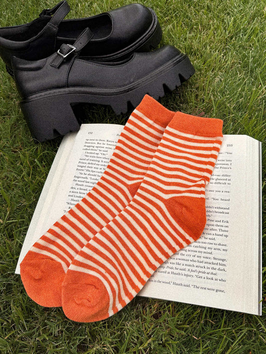 Rust orange and ivory striped crew socks named The Life of a Cozy Girl Socks placed on an open book next to black platform shoes on grass.