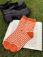Rust orange and ivory striped crew socks named The Life of a Cozy Girl Socks placed on an open book next to black platform shoes on grass.