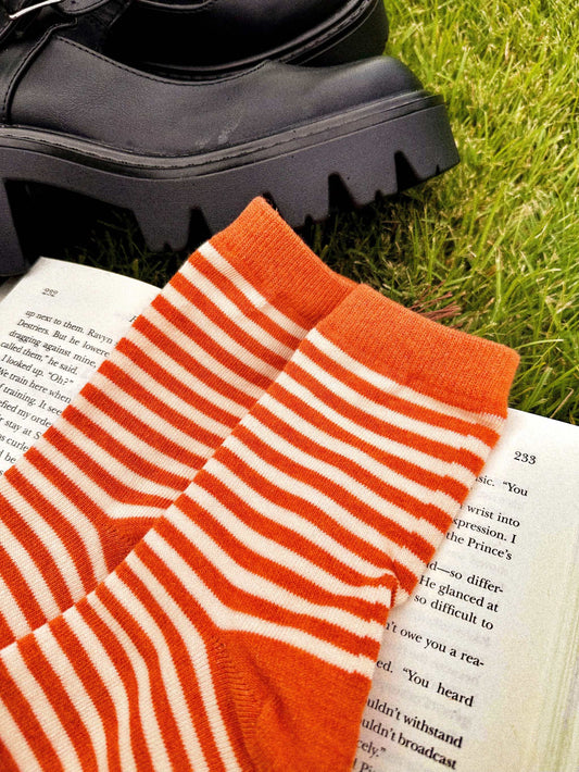 Striped rust orange and ivory crew socks resting on an open book next to black boots representing The Life of a Cozy Girl Socks.