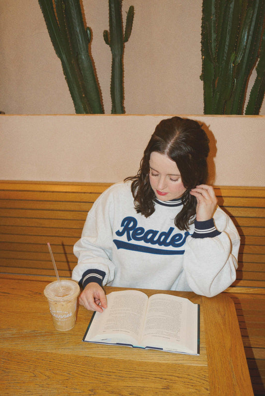 Woman wearing ash gray Reader Crewneck with navy striped trim reading a book at wooden table with iced coffee