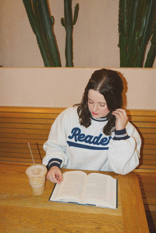 Woman wearing ash gray Reader Crewneck with navy striped trim reading a book at wooden table with iced coffee