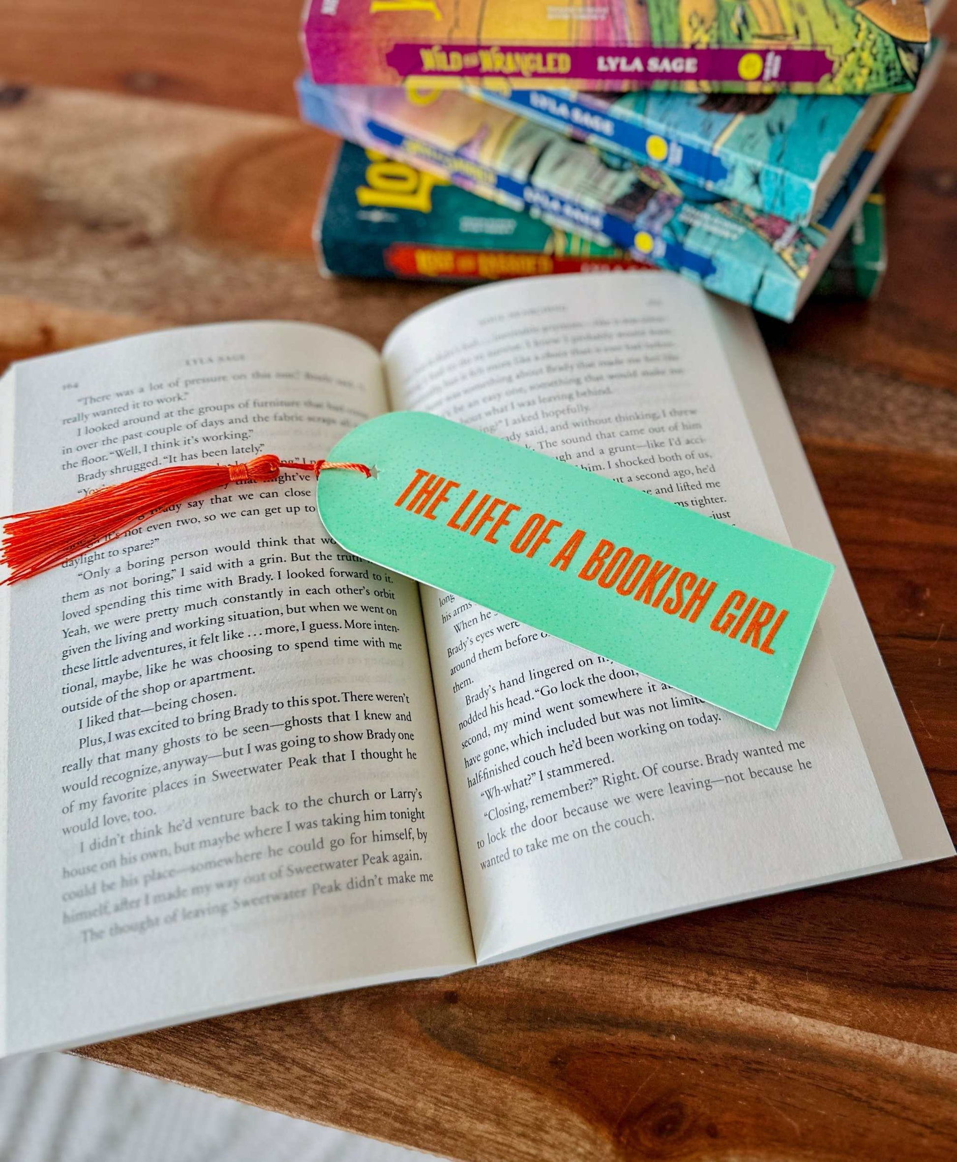 Open book with a bookmark titled 'The Life of a Bookish Girl' on a wooden surface with other books stacked in the background. Created for the bookish swifties to celebrate The Life of a Showgirl.