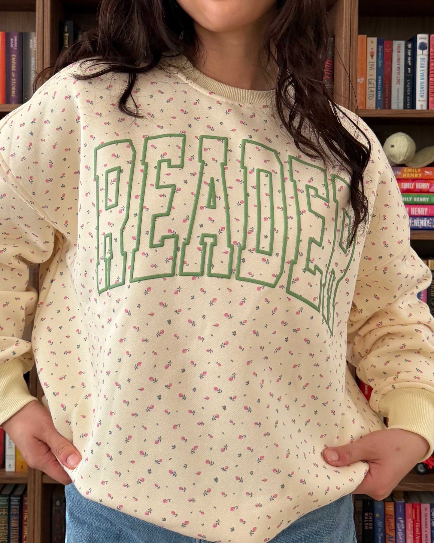 The Reader Floral Crewneck from Next Chapter has a pale yellow fabric with a subtle floral pattern. Across the sweatshirt is the word 'READER' screen printed in green. Next Chapter offers high quality bookish merch and apparel. 