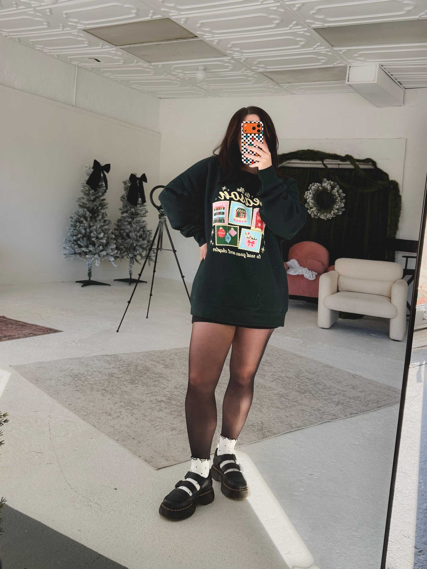 Woman wearing a festive Next Chapter Holiday Crewneck Sweatshirt with bookish Christmas-themed graphics, holding a phone, standing indoors.
