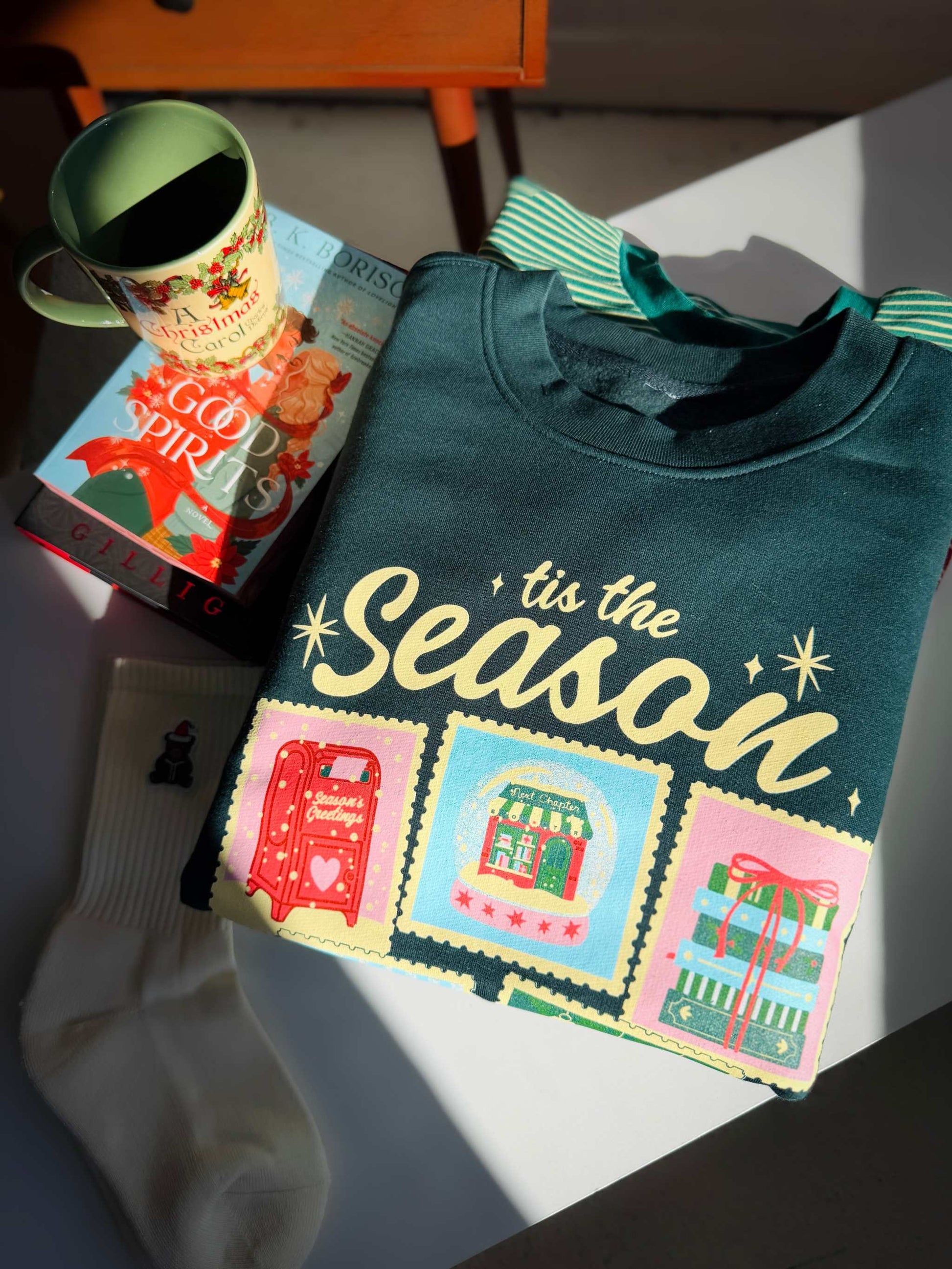 Next Chapter Holiday Crewneck sweatshirt with 'tis the Season' text and festive bookish Christmas graphics on a table with a mug and box in the background.