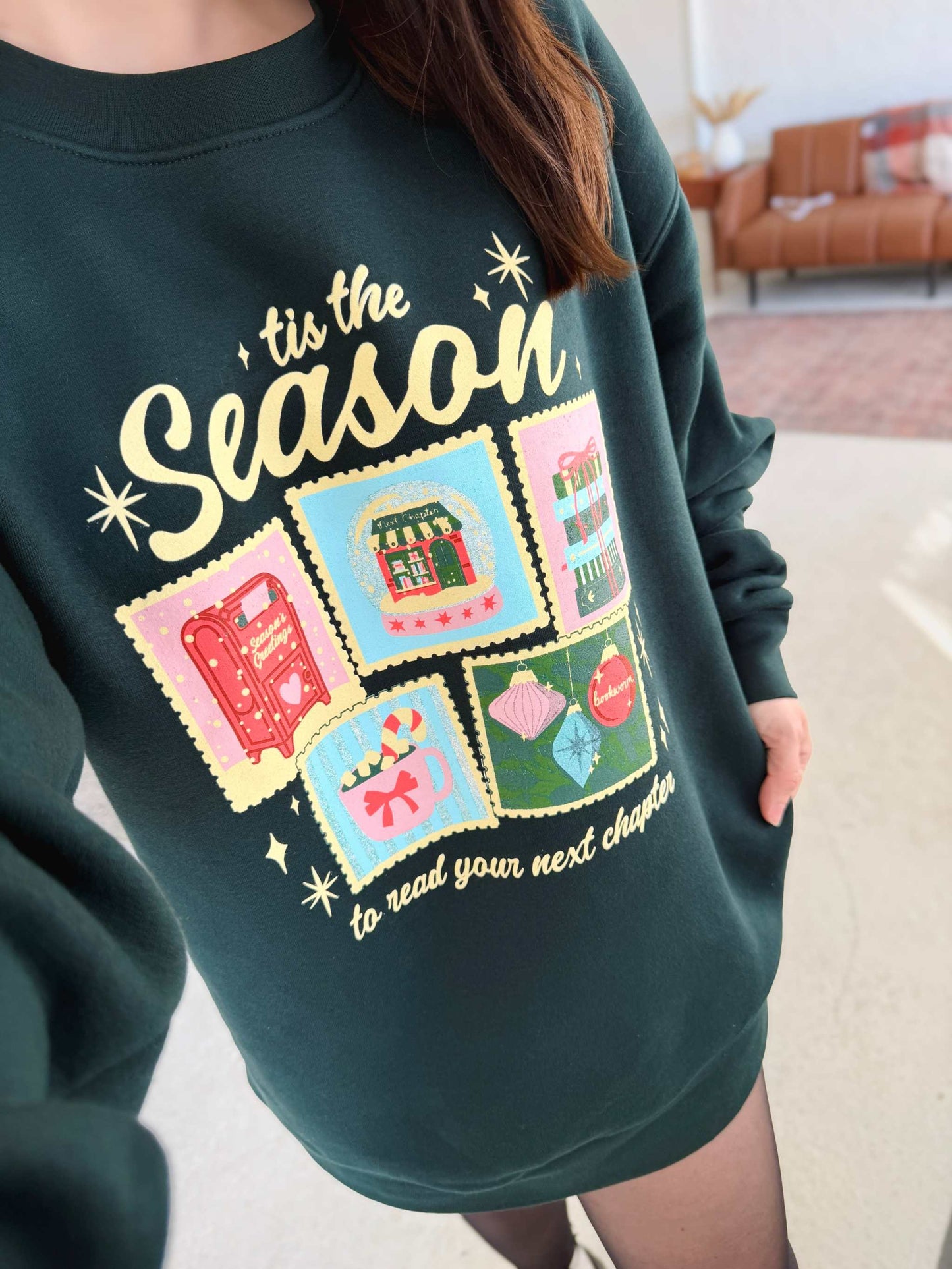 Next Chapter Holiday Crewneck. A Dark green sweatshirt with a festive bookish Christmas design and 'tis the season' text in a casual indoor setting.